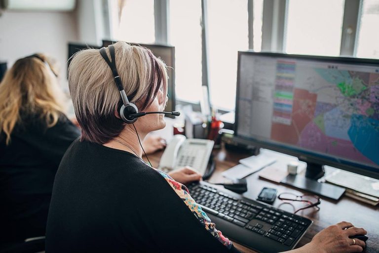 A person wearing a headset sits at a desk with two monitors displaying maps and data. They are typing on a keyboard. Another person is visible in the background, also wearing a headset and working at a computer.
