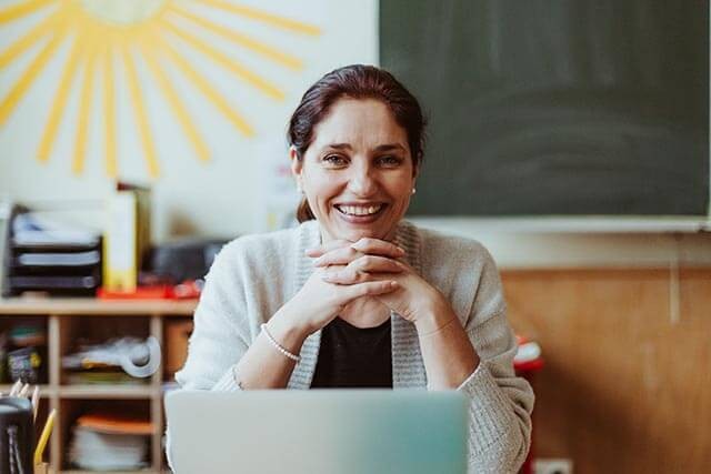 A woman sitting in a desk in a classroom, smiling at the camera. Her hands are clasped in front of her.