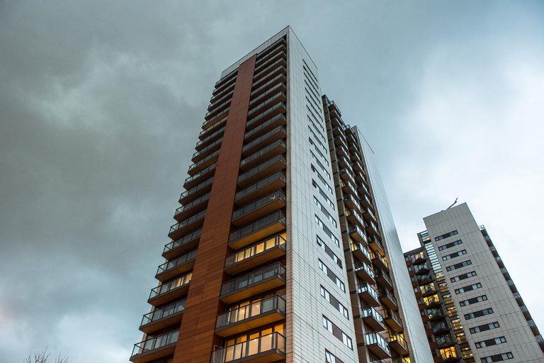 Tall apartment building with balconies, cloudy sky, and illuminated windows. Adjacent to it is another tall building.