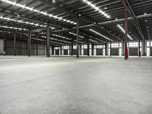 Spacious, empty warehouse with concrete floor, metal beams, and natural light coming in from big windows.