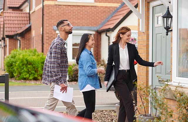 Estate agent opening a door to show a house to a couple in a residential area.