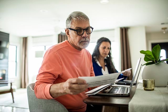 An older man in glasses is sitting at a table and checking some documents. In the background, a woman is also checking some documents.