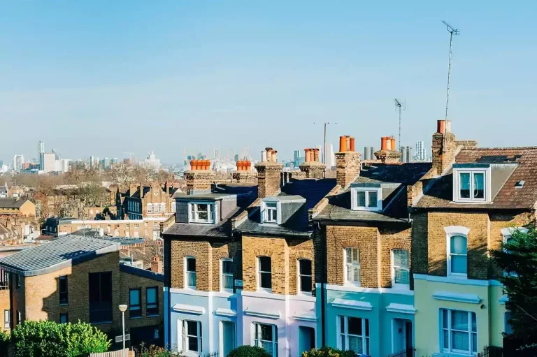 Row of brick traditional terraced houses on a residential street with chimneys and tall buildings visible in the background.