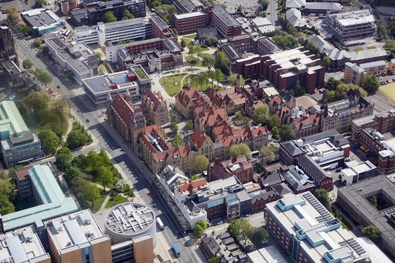 Aerial view of a university campus with red-brick and modern buildings, surrounded by trees and paths.