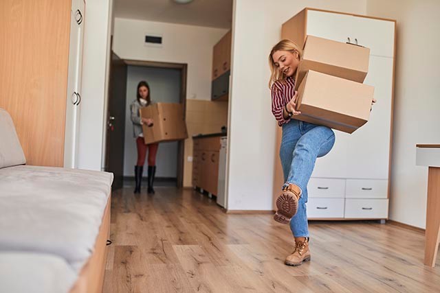 Two students moving boxes in an empty room with wooden floors and simple furnishings.