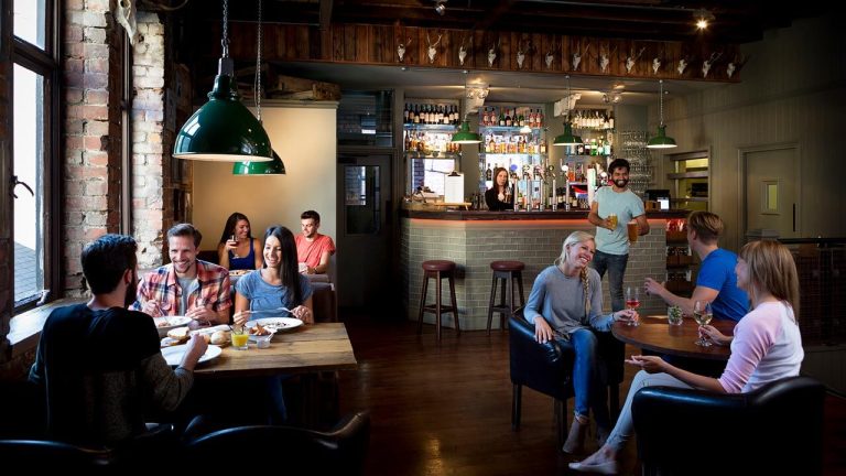 Cozy restaurant with wooden tables, groups dining, and a bartender serving drinks in a warm, rustic setting.
