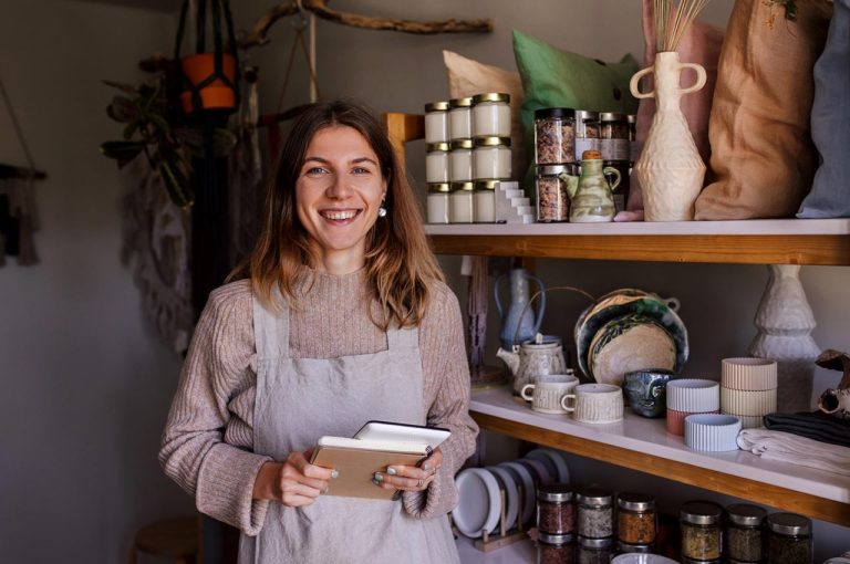 Person smiling and wearing an apron in holding an notebook stands in a cozy store filled with jars, and pottery products.