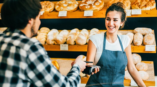 A customer is pointing a card to make a payment in a bakery. Bakery goods are on the shelves in the background.