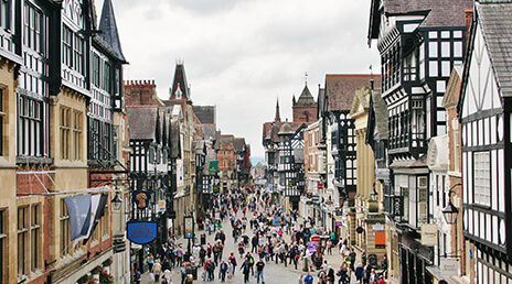 A bustling street scene in a historic town, featuring Tudor-style buildings with black and white timber framing. The street is filled with people walking, and the sky is overcast, adding a subdued atmosphere to the scene.