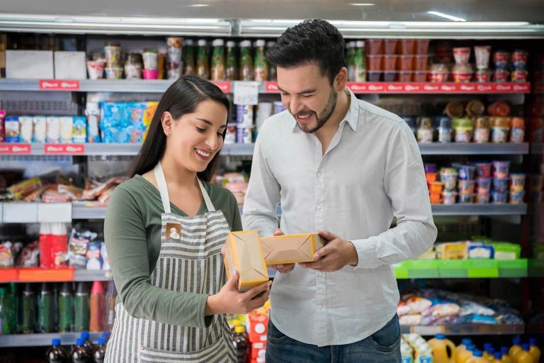 A man and a woman stand in a grocery store aisle, smiling and holding food boxes.