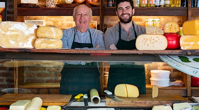Two deli workers stand behind a cheese counter. Various cheese wheels and wedges are displayed before them.