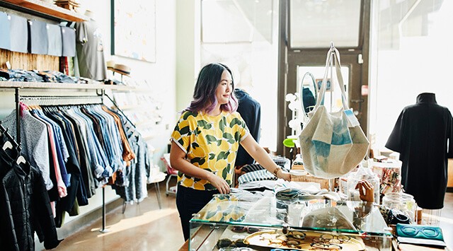 A woman with purple-tinted hair is browsing inside a clothing store. She is wearing a yellow top with a leaf pattern. The store displays various clothes on racks and accessories on a glass counter. Natural light streams in from large windows.