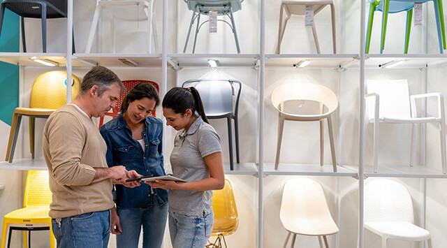 Three people in a furniture shop are standing by a shelving unit with chairs, looking at a tablet that one of them is holding.