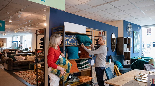 Man and woman selecting cushions in a furniture store, surrounded by couches and decor items.