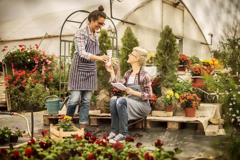 Two people in a greenhouse, one handing a cup to the other seated with a notebook in her hand.