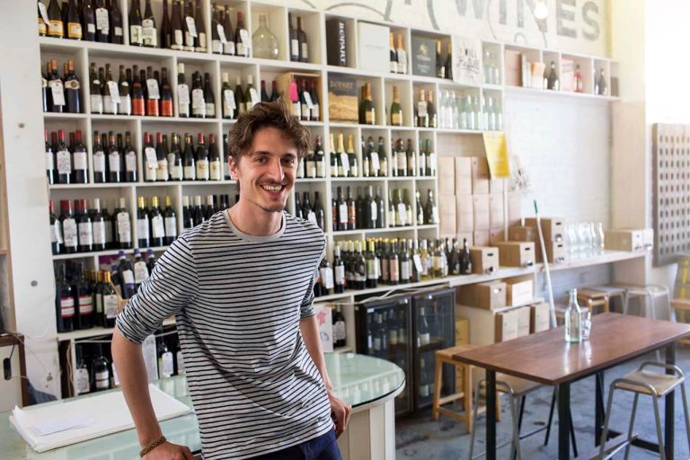 A person smiles while standing in front of a counter of a wine shop and shelves filled with various wine bottles.