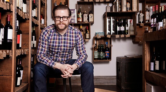 A man wearing glasses and a plaid shirt sits on a stool in a wine cellar, an off licence treasure trove behind him. Shelves brimming with bottles of wine and spirits frame his neutral expression as he gazes at the camera.