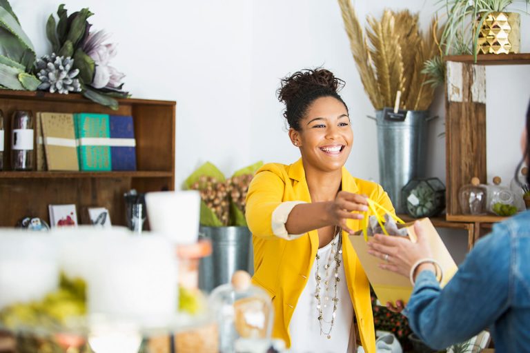 Smiling store owner handing a shopping bag to a customer in a cozy shop with plants and various items.