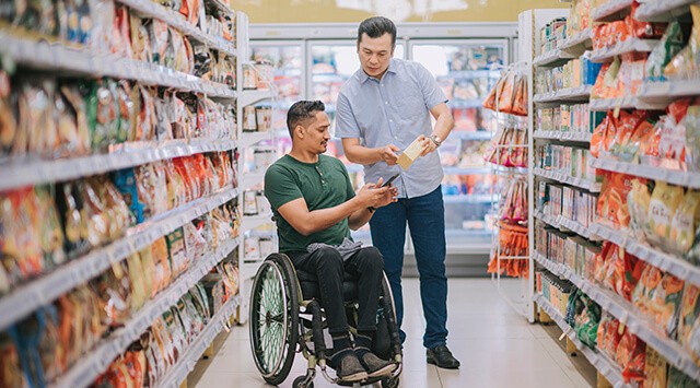 Man in a wheelchair in a grocery aisle, assisted by another man holding a box.