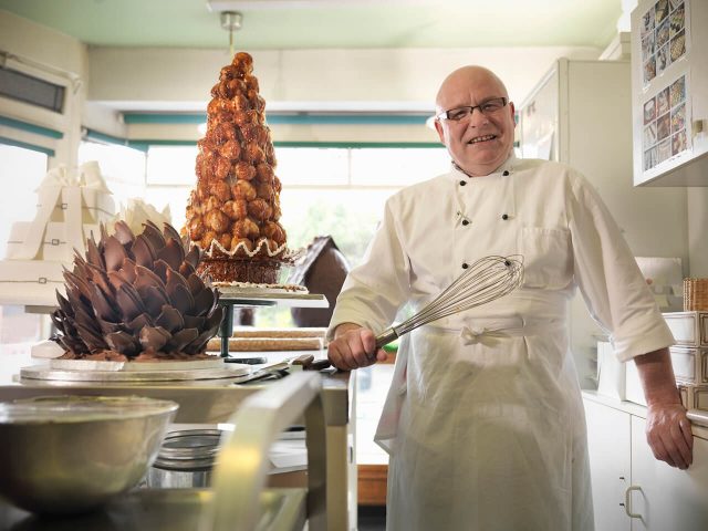 Chef holding a whisk, with a croquembouche and chocolate sculpture on the counter.