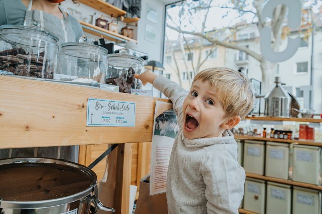 Child with an astonished expression points at chocolate jars in a sweet shop.