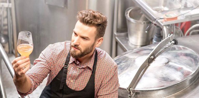 Man with a beard wearing a checkered shirt and apron holds a glass of beer, examining its color. He is in a brewery, surrounded by brewing equipment, including a large metal container with a sealed lid in the background.