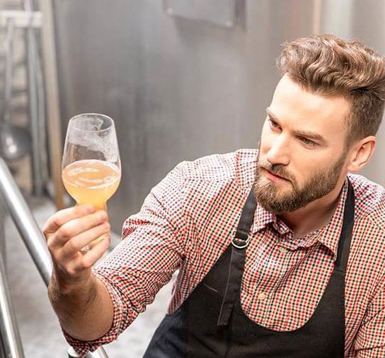 A man in a checkered shirt and apron examines a glass with a light-colored liquid, possibly beer, in an industrial setting. He has a beard and is focused on the contents of the glass.