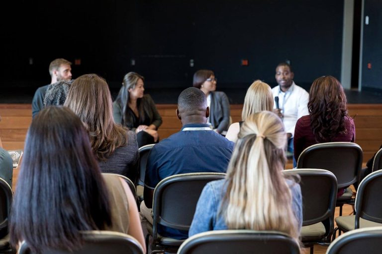 Audience seated and facing a panel of four speakers on stage.