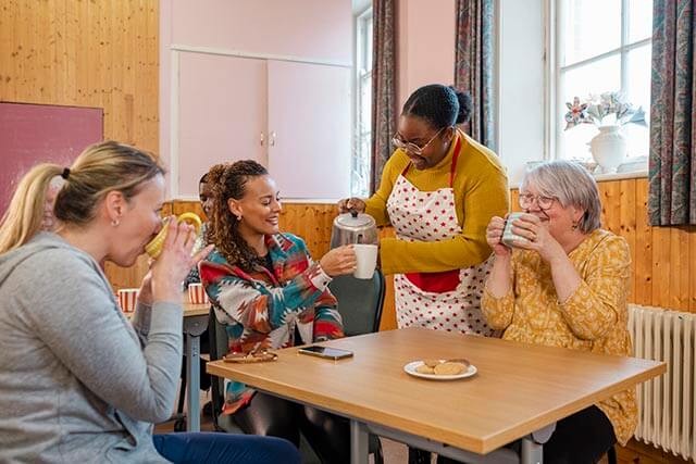 Three women are enjoying tea and biscuits in a bright sports community centre. A waiter pours tea into a mug.
