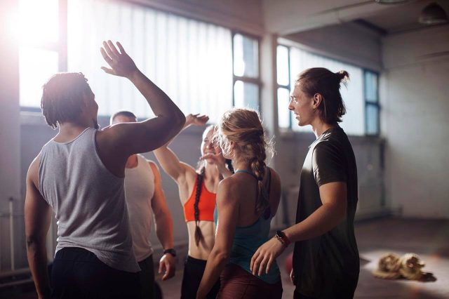 Group of five people in athletic wear smiling and giving high-fives in a sunlit gym.