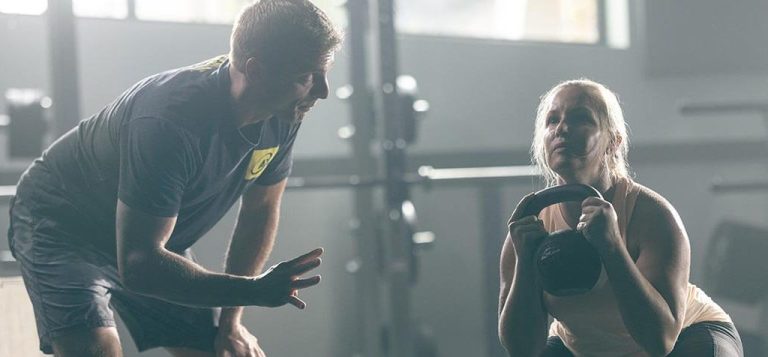 Female client doing a kettlebell squat in a gym, with a man offering guidance.