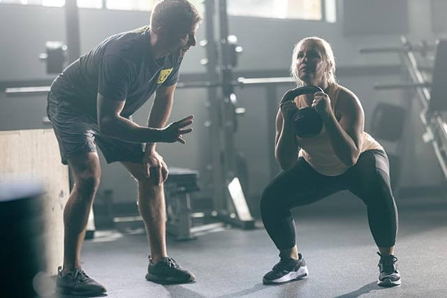 A woman executes a squat with a kettlebell in a well-equipped gym. Beside her, a man crouches, providing guidance. With weights and machines around, the gyms atmosphere highlights the need for proper training and gym insurance to ensure safety for all.