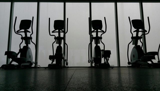 Four elliptical machines lined up in a dimly lit gym. Only the shape of the equipment is visible.