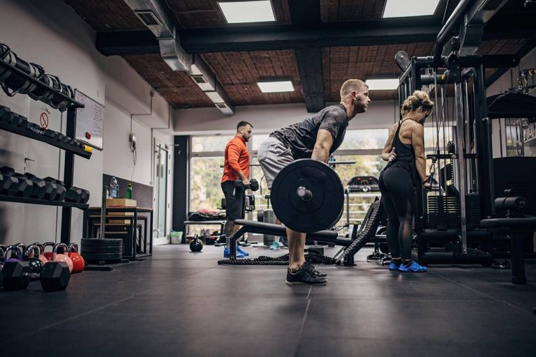 Man lifting barbell in a gym while others work out, surrounded by gym equipment.