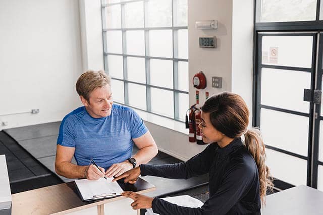 Man reviewing gym forms while chatting with a woman in a bright room with large windows.