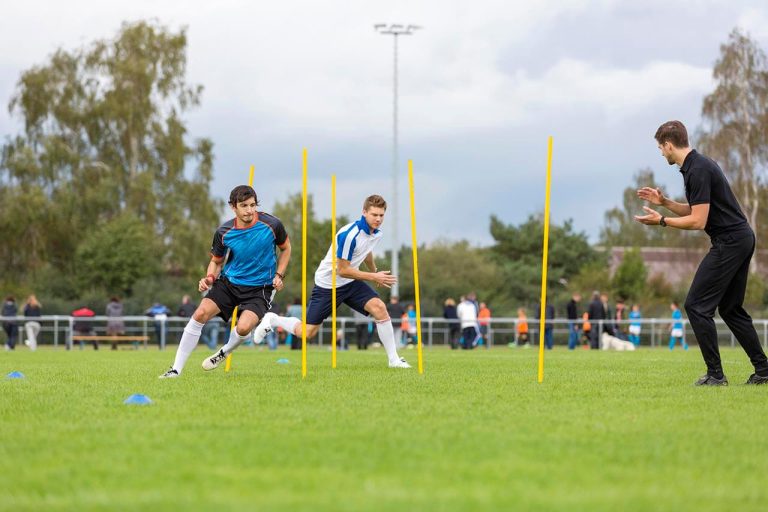 Football players running between poles for agility practice, with a coach clapping nearby.