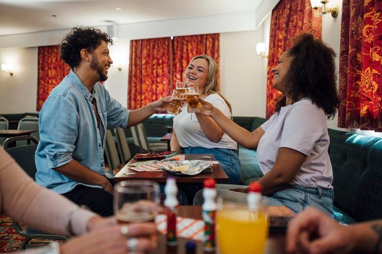 Three people clinking glasses in a social club, with drinks and snacks on the table and bingo cards.