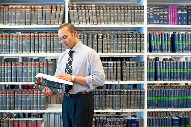 A man dressed in shirt and a tie in front of bookshelves, holding an open book and focused on reading.