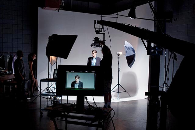 A person stands in front of a white backdrop, posing for a photoshoot. Studio lights, umbrellas, and other equipment surround them. A monitor displays the persons image, and silhouettes of crew members are visible.