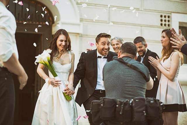 A newlywed couple stands outside a building, surrounded by smiling guests. The bride holds a bouquet of white flowers and wears a white dress, while the groom is in a black suit. Flower petals are being tossed in celebration. A photographer faces them.
