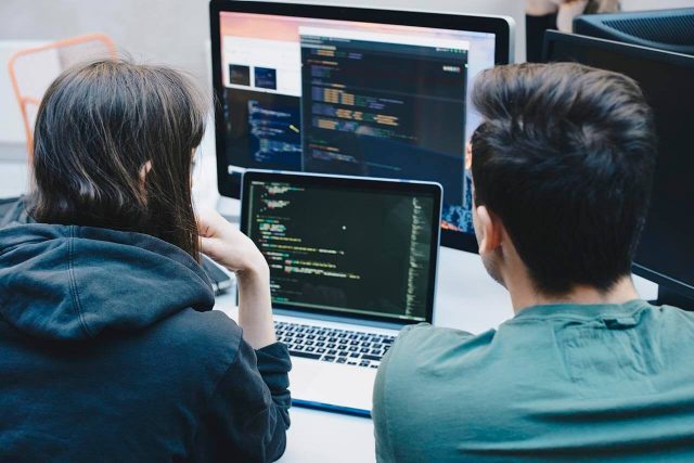 Two people sitting in front of computer screens displaying code. The person on the left is wearing a dark hoodie, and the person on the right is wearing a green shirt. They are engaged in programming or debugging in a workspace.