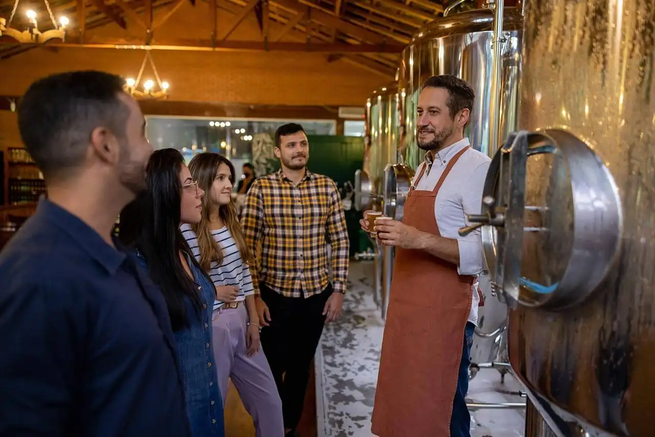 Group of five people on a guided tour inside a brewery, standing near large stainless steel brewing tanks.