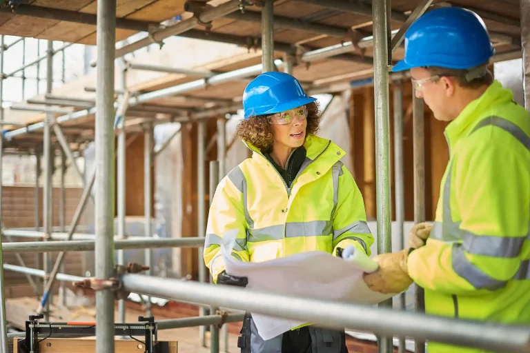 Construction workers in yellow jackets discussing plans at a site with scaffolding.