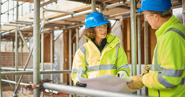 Two construction workers in yellow jackets and blue helmets discuss plans at a site covered by scaffolding.
