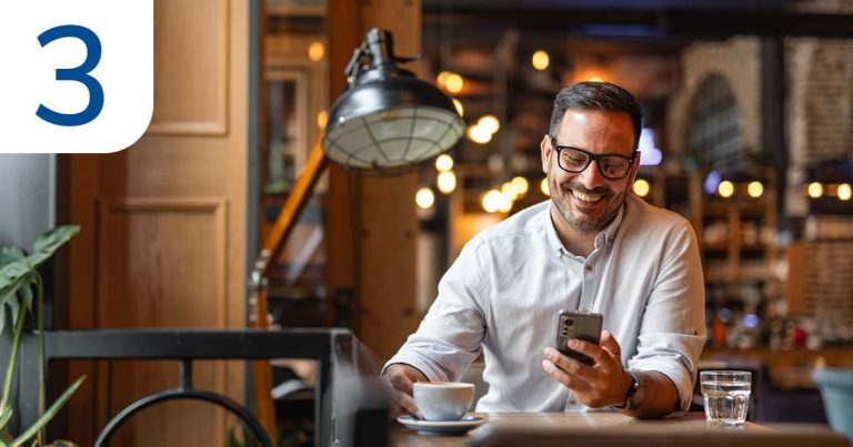 A content business owner wearing glasses sits at a table, engaged with his phone and smiling.