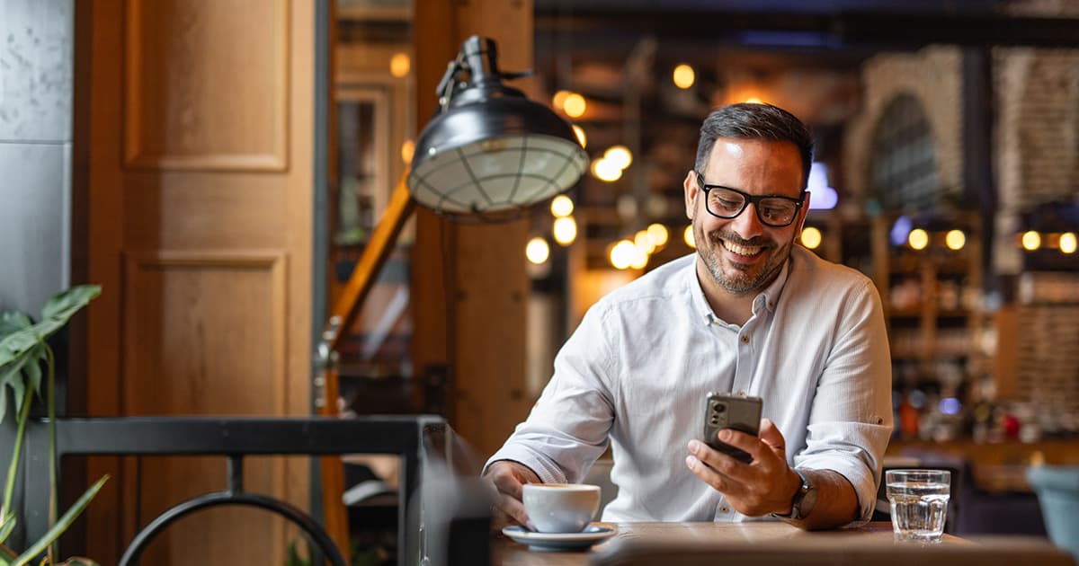 A content business owner wearing glasses sits at a table, engaged with his phone and smiling.