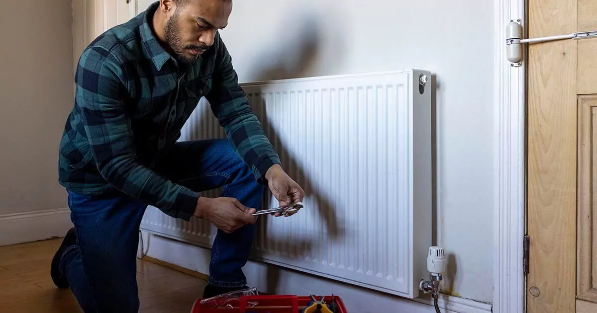 A man is fixing a radiator in a room, surrounded by tools, concentrating on the maintenance task at hand.