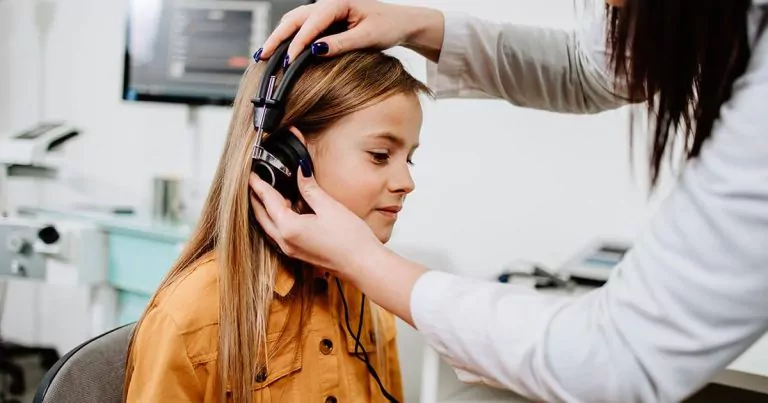 Young girl sitting while a woman adjusts headphones in an audiology office.