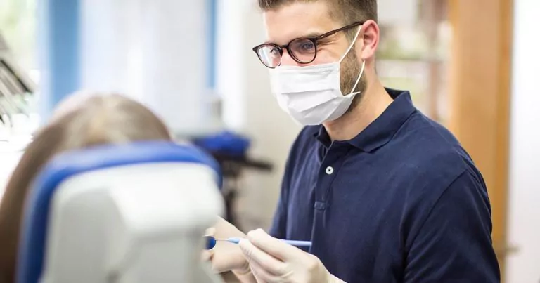 Dentist in navy shirt and mask focused on a patient sitting on a dentist chair.