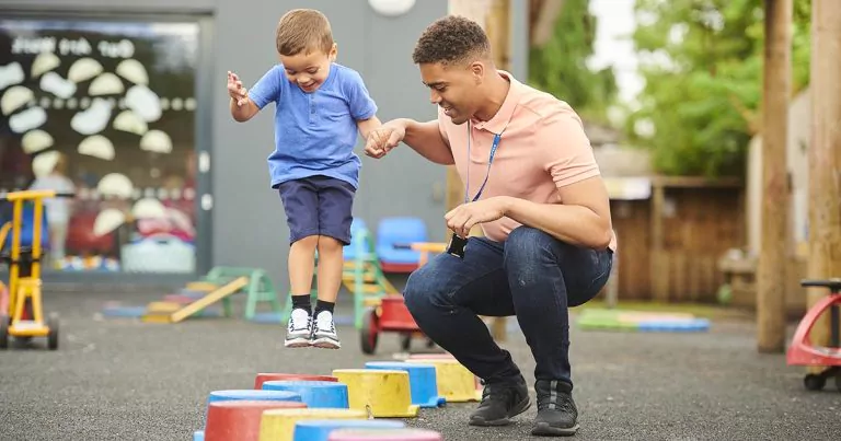 Nursery worker helping a child step over colorful playground stones.